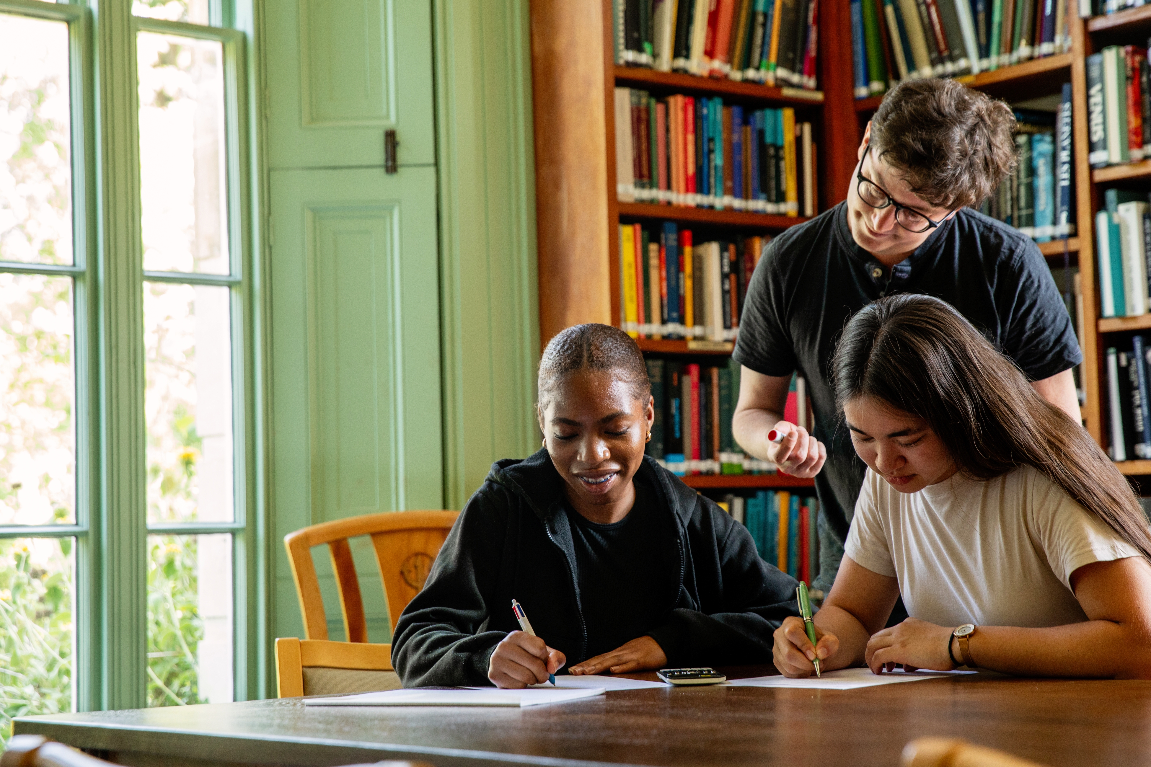 Two students sitting writing, whilst a third person looks over their shoulder.
