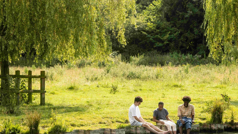 Three students chatting and sitting in a field with their feet in the river.